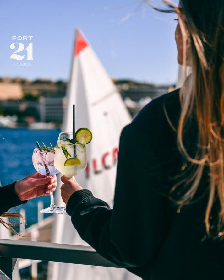 Two gin and tonics raised by the water with a sail behind